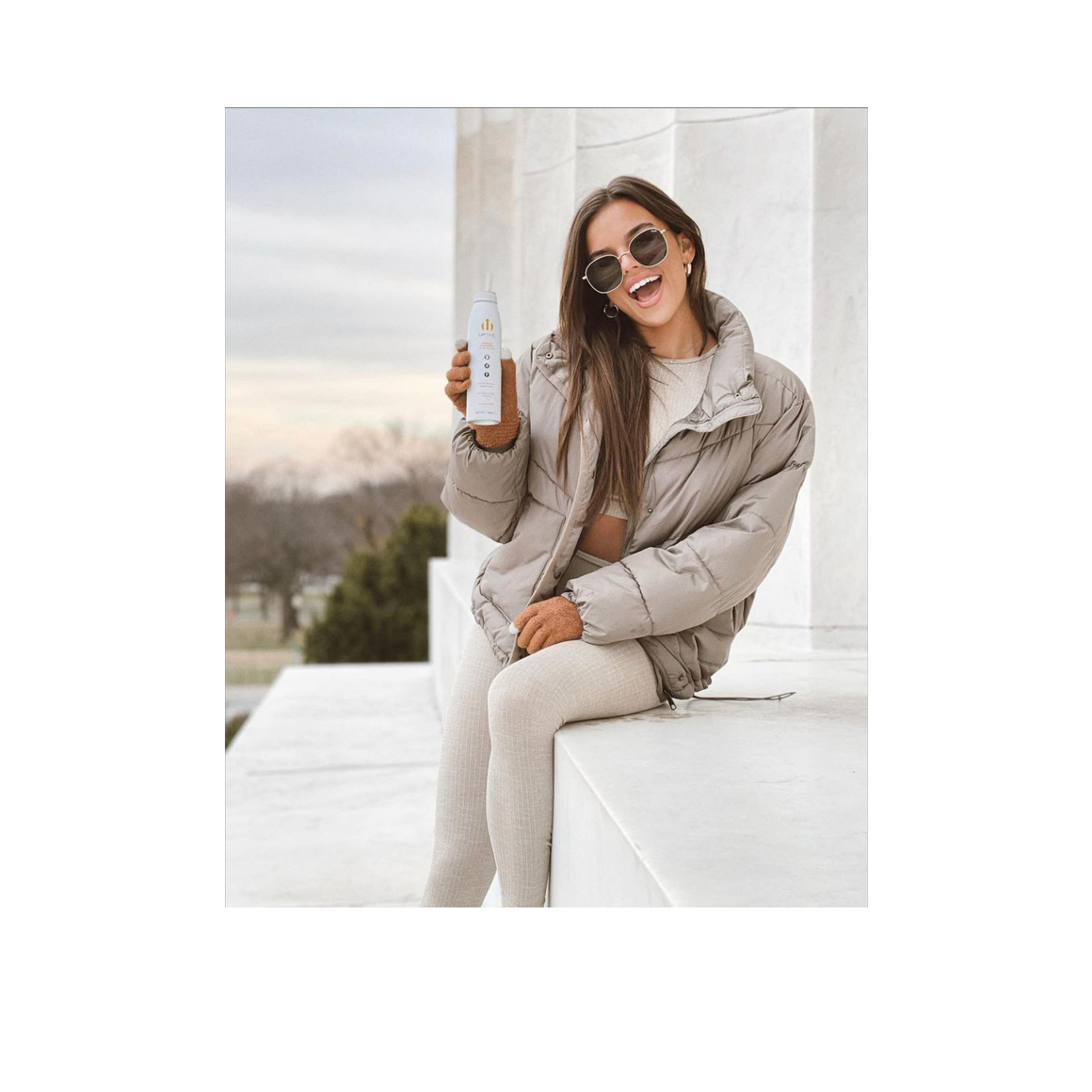 smiling woman in sunglasses sitting on a step holding a bottle of uptime mango pineapple energy drink Image3